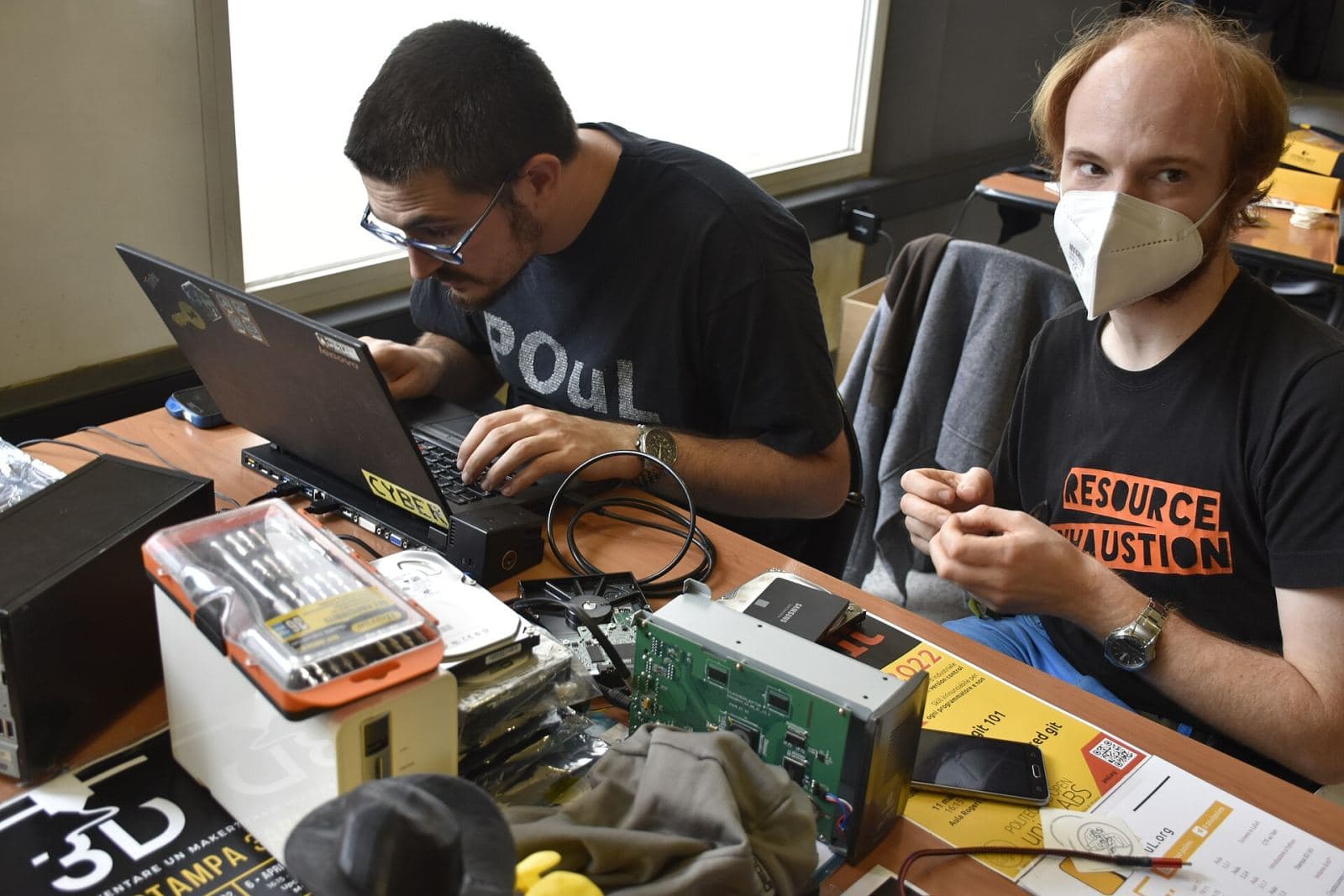 Technicians working at a computer repair workshop bench surrounded by laptops, disk drives and repair kits